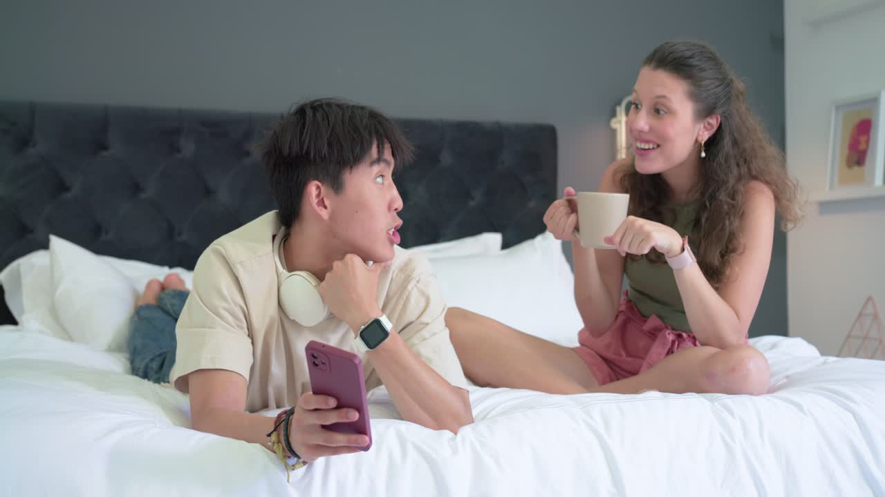 Diverse couple chatting on bed, woman pointing at pink smartphone, man showing screen to share