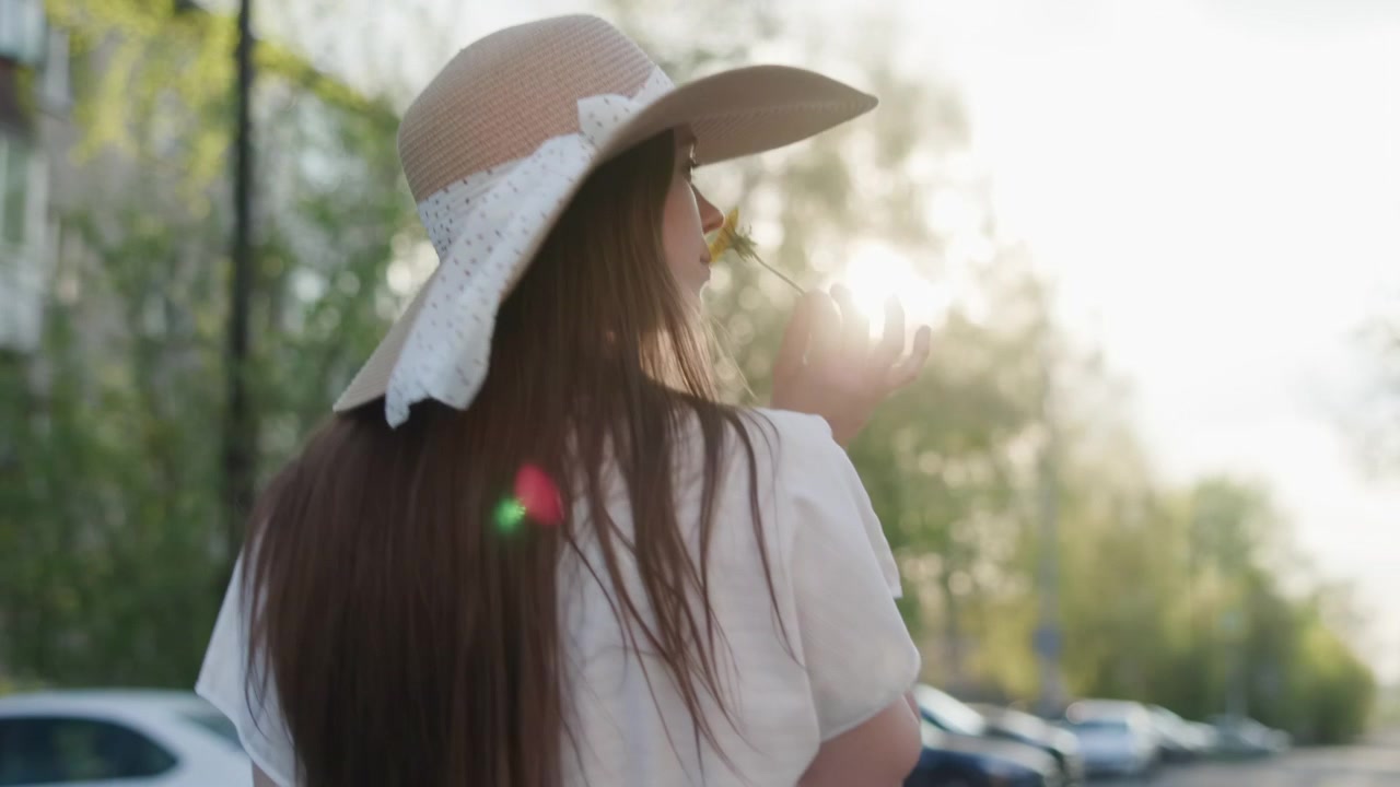 Rear view of a woman in a straw sun hat and white top strolling through a peaceful neighborhood with parked cars, green trees, and warm evening light