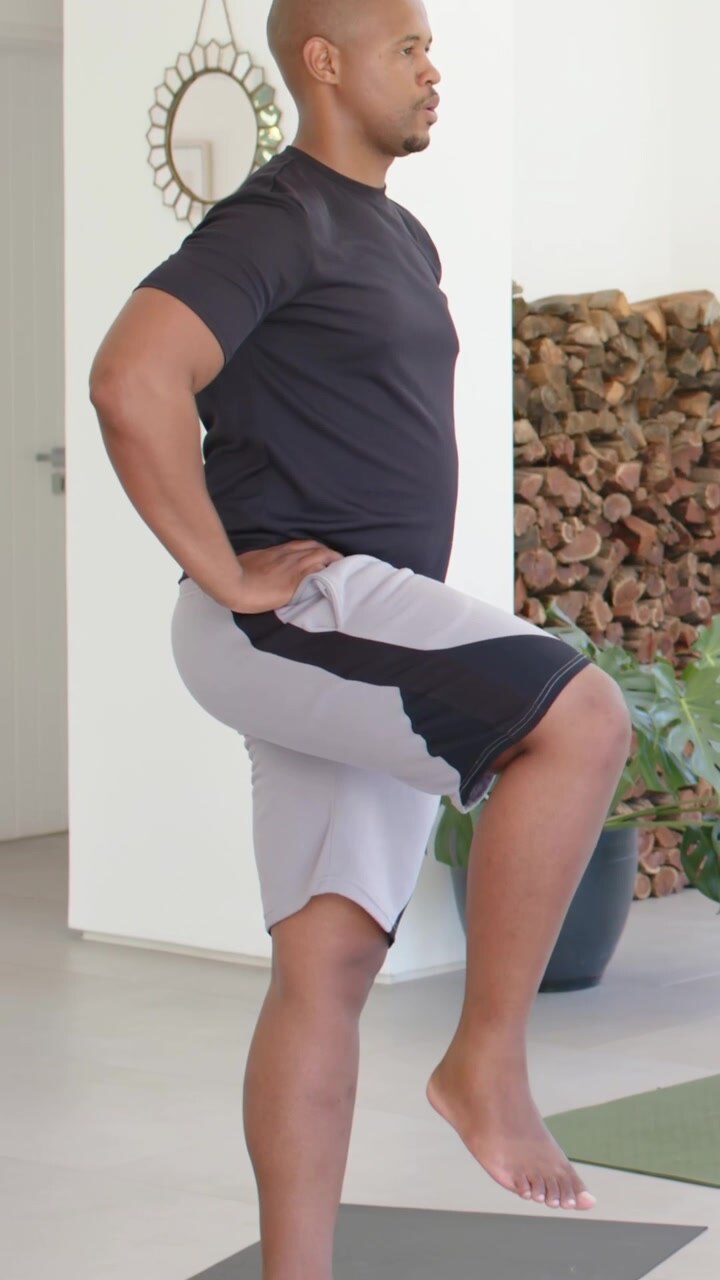 Vertical video: Starting man lifting right knee at home for balance in tee shorts near mat plant