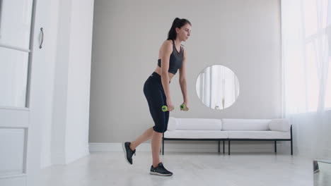 Young-beautiful-brunette-woman-standing-on-one-leg-doing-bends-with-dumbbells.-Performing-deadlift-with-dumbbells-at-home.