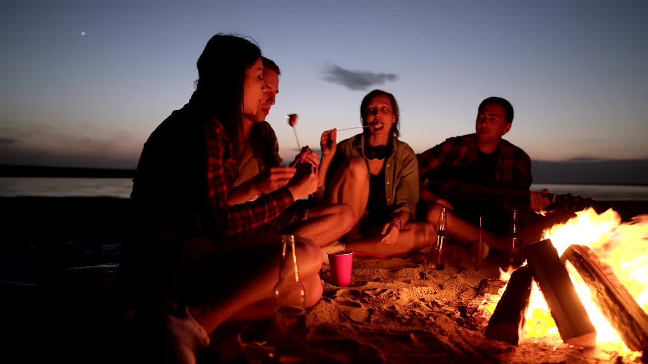 해변에 앉아있는 친구들의 가속화된 영상, 불꽃 근처의 플레이드, 나무 막대기로 음식을 기, 먹기, 대화, 행복한 시간, 우정, 밤, 바람이 불고 있습니다.