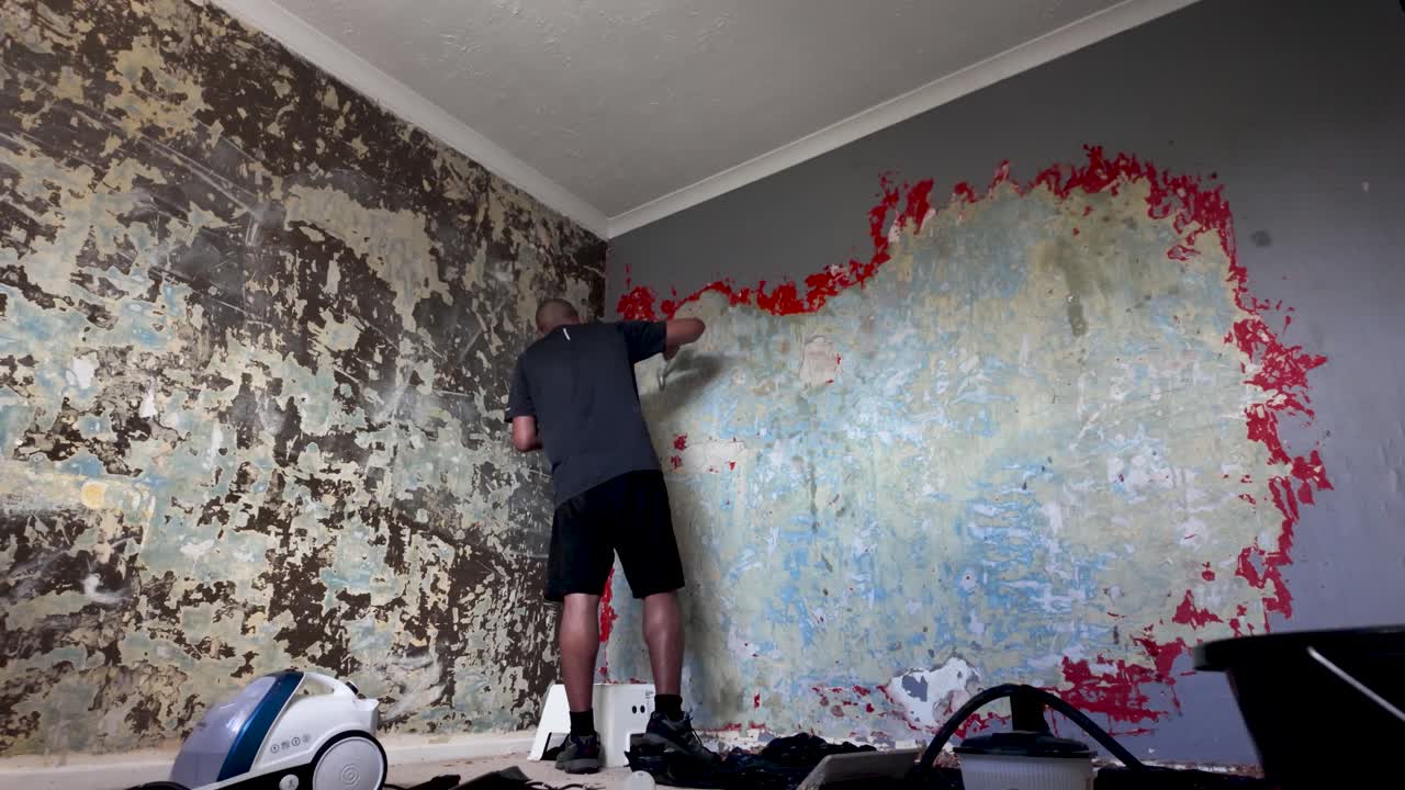 A handyman in shorts removes peeling wallpaper in a dim, aged room during renovation project