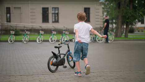 a young boy is seen from behind, running excitedly toward a parked black bicycle, in the background, several bicycles are lined up, with a man standing nearby and another child riding a bike