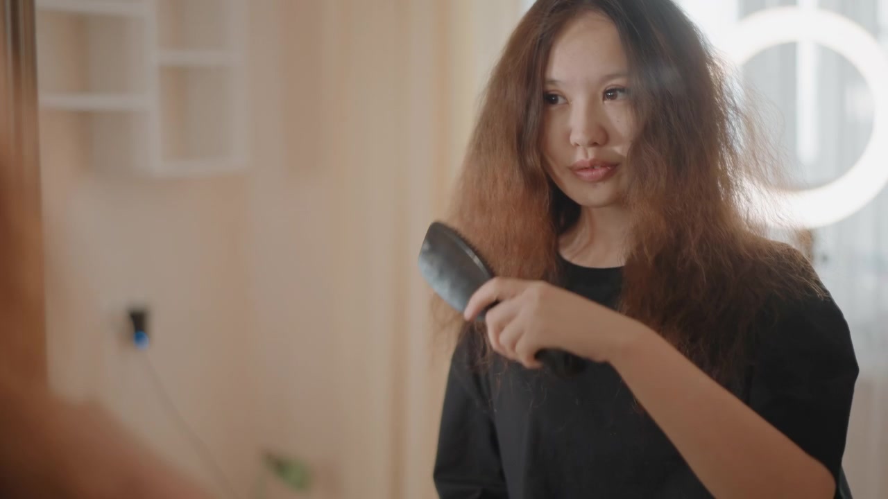 Mujer joven con cabello largo y rizado de pie de cintura para arriba frente al espejo, hablando y gesticulando mientras se cepilla el cabello, con contraluz suave, reflejos en el contorno, tonos cálidos y una atmósfera de estilo de vida tranquila.