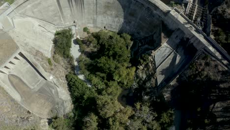 Luftaufnahme,-Die-Gerade-Nach-Unten-Schaut-Und-über-Einen-Großen-Gekrümmten-Betondamm-Fliegt,-Gleitet-über-Einen-Dunklen-Glasigen-Stausee-Und-Ein-Kleines-Fahrendes-Boot-Hinter-Dem-Damm