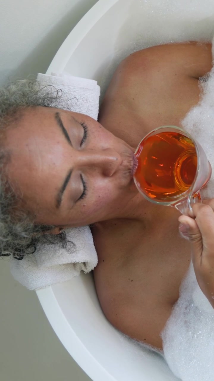 Vídeo vertical: Mulher idosa levando copo de vidro, tomando bebida na banheira, descansando com toalha, espuma.
