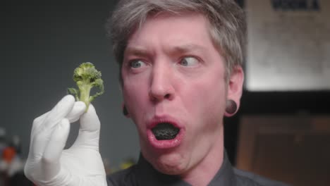 young crazy male chef in an elegant black shirt with an alternative look, tattoos holding mushrooms, looking excessively at broccoli and spitting out a piece