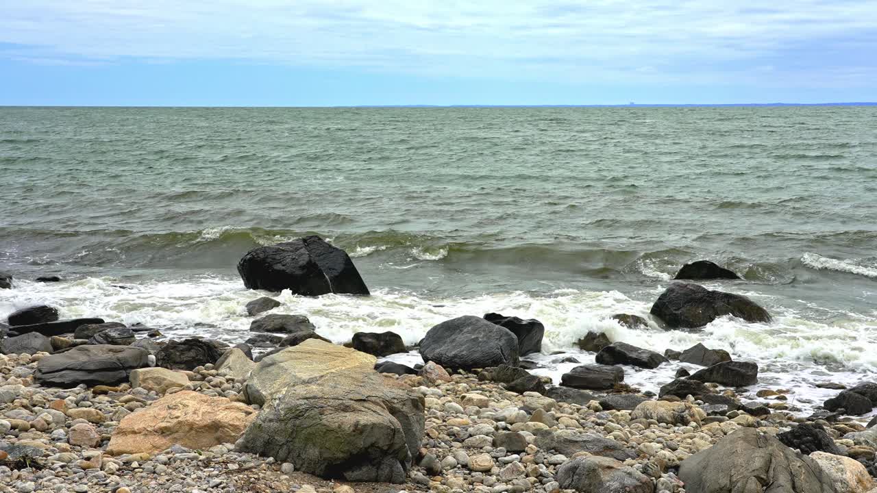 Enjoy the calming sounds as waves crash against a rocky shore during daylight. The soft clouds above create a peaceful atmosphere by the water