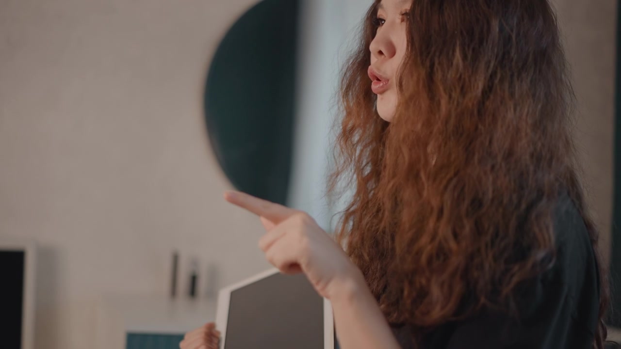 Vista de perfil de una mujer, de pecho hacia arriba, sosteniendo una tableta de pantalla oscura en una mano y señalando con el dedo índice; una ligera inclinación hacia adelante y una expresión concentrada sugieren interacción, explicación o guía
