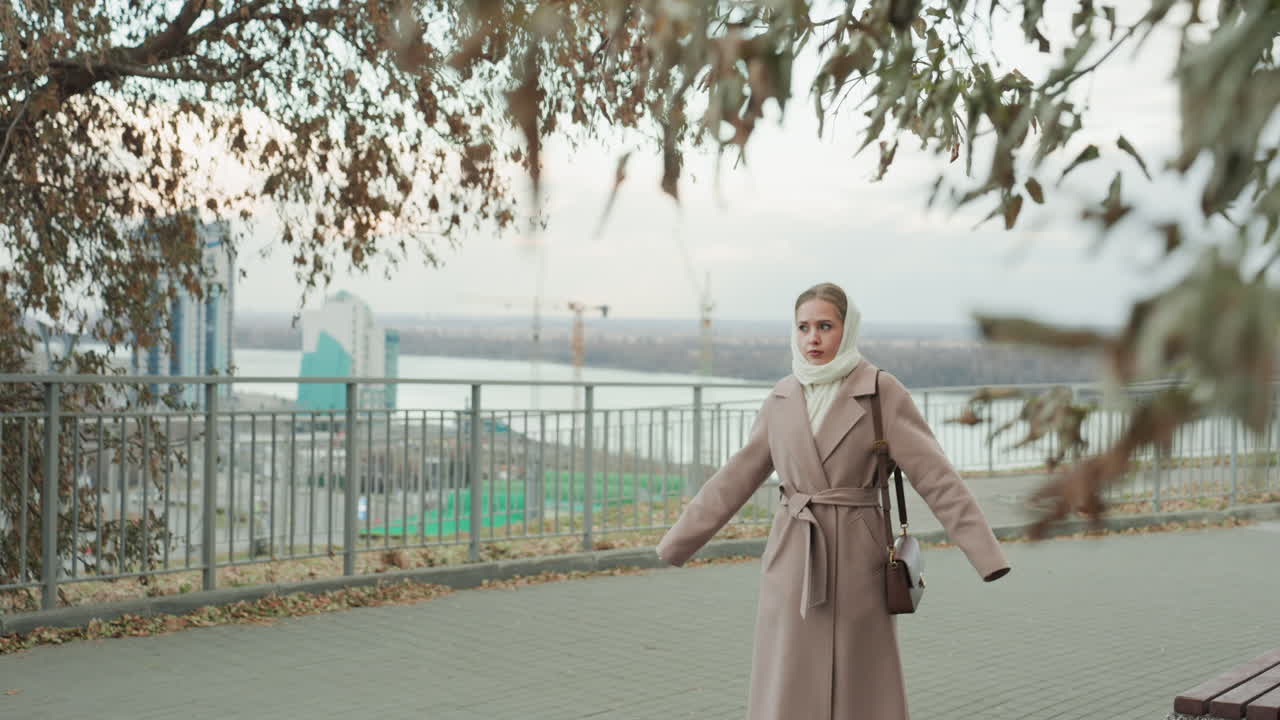 Donna bianca che cammina sul lungomare in autunno, indossa un cappotto lungo e una sciarpa color crema, mani giunte, cielo coperto e tenue, foglie che cadono, skyline della città in lontananza, sguardo contemplativo, brezza leggera.