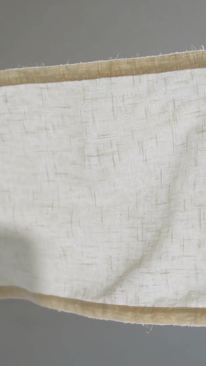 Vertical video: Breeze lifting beige woven cloth unfurling across gray wall, showing stitched hem