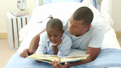 afroamerican little boy reading a book with his father in bed