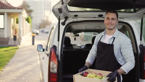 hombre amigable sonriente tomando de la caja de madera del baúl con manzanas en la calle al aire libre, retrato de hombre de entrega feliz en delantal