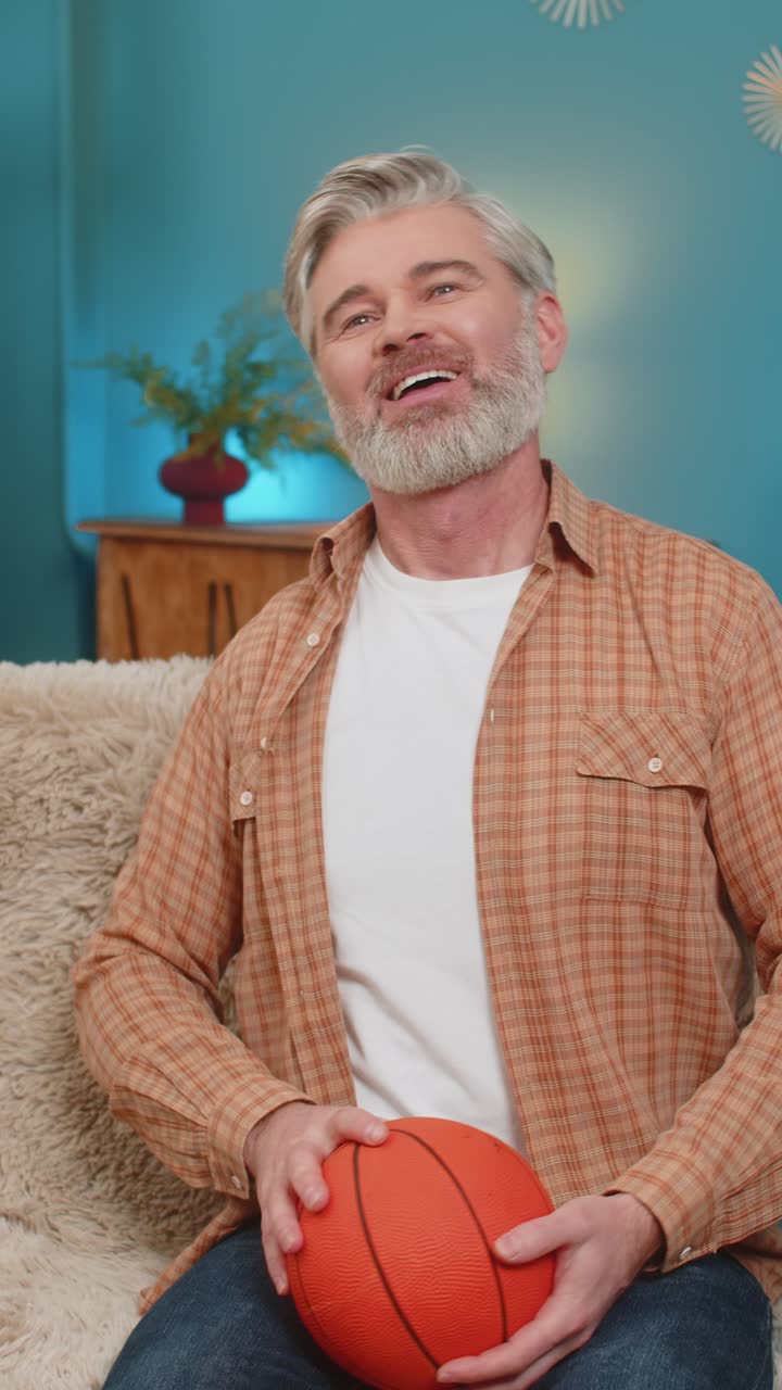 Mature man sitting on sofa with basketball tossing and spinning ball enjoying playing at home
