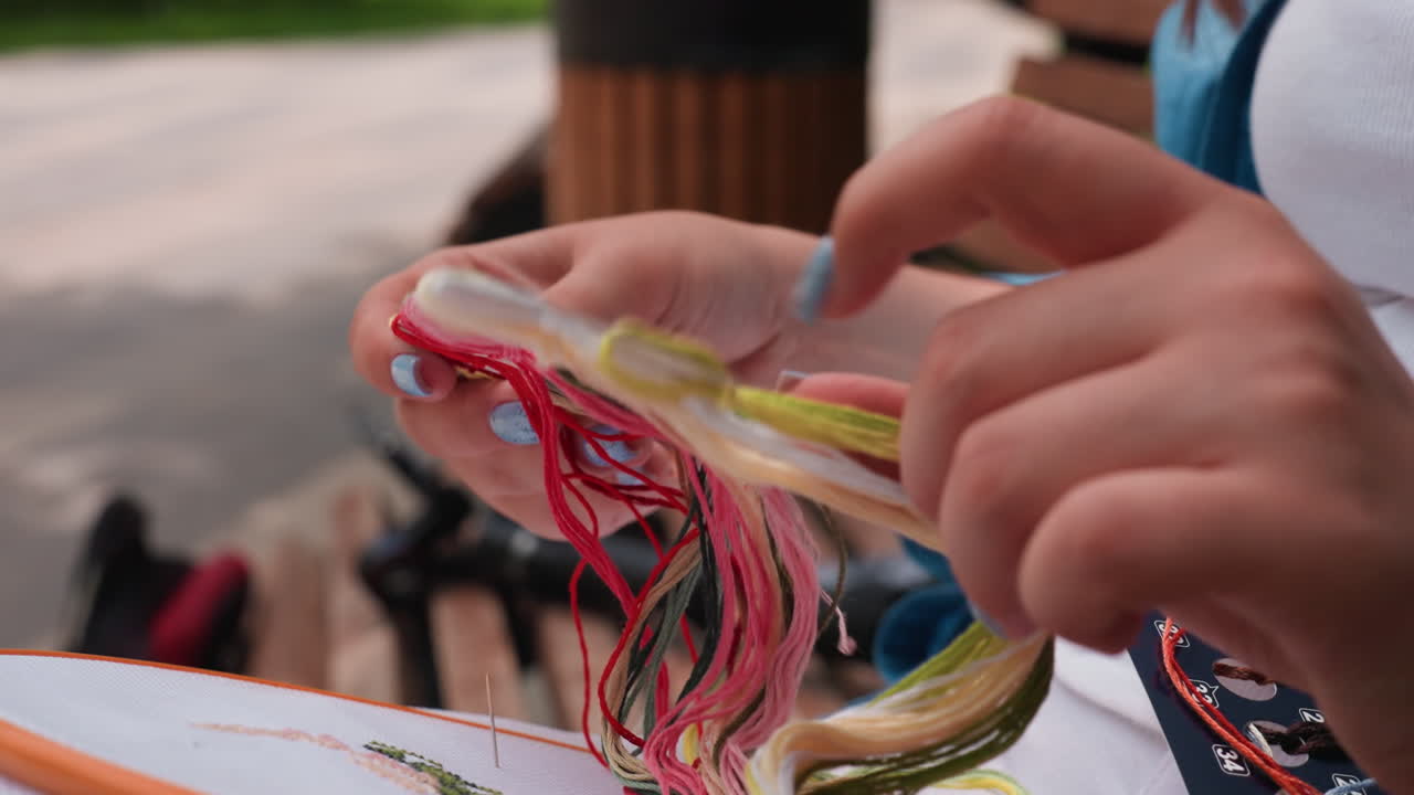 Close up hands separating tangled embroidery thread from multicolor floss card, blue nails choosing green strand beside hoop and stitched fabric, careful motion resolving knots