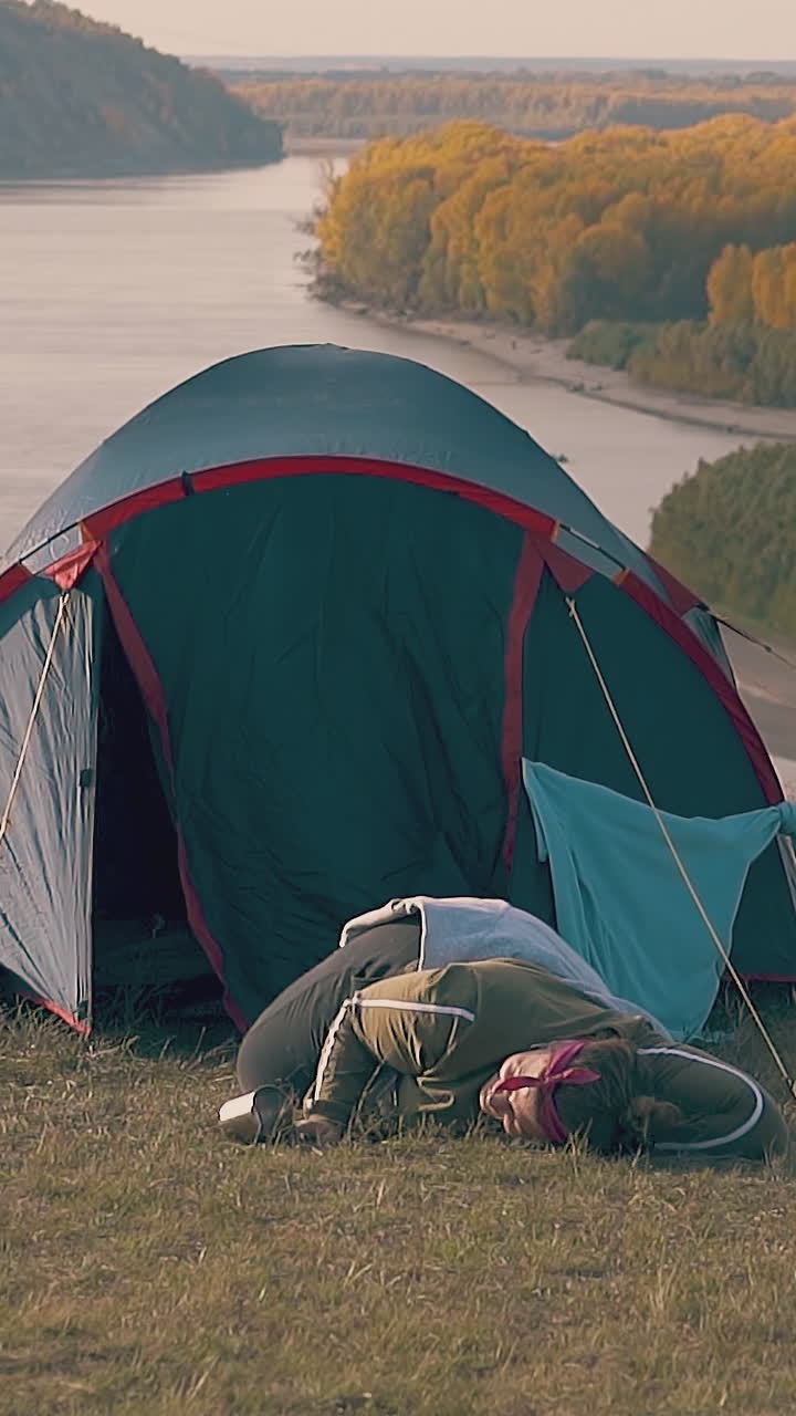 pudgy girl with hard hangover lies at blue tent holding metal cup against nice river in early autumn morning slow motion