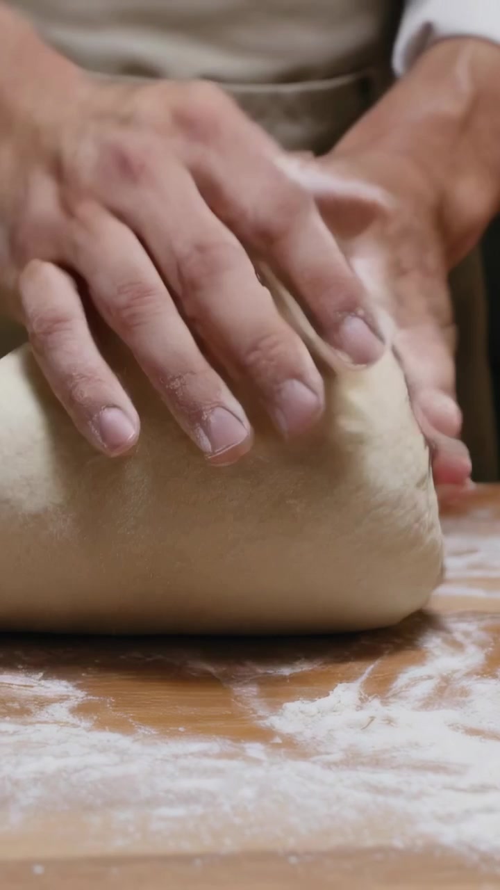 Video vertical: Manos de cocinero iniciando el pliegue para formar una hogaza de levadura sobre una encimera enharinada para hornear, con delantal.