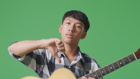 close up of asian teenager with guitar showing thumbs down gesture while standing on green screen background