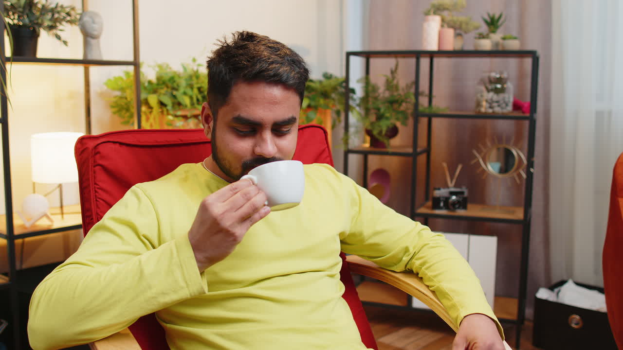 매력적인 미소 짓는 인도인 남자가 아침에 집 의자에서 커피 한 잔이나 허브 차를 마시고 있습니다.
