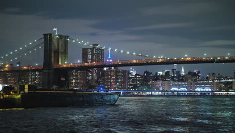 cargo barge sails under the brooklyn bridge in new york on the background of the night lights of man