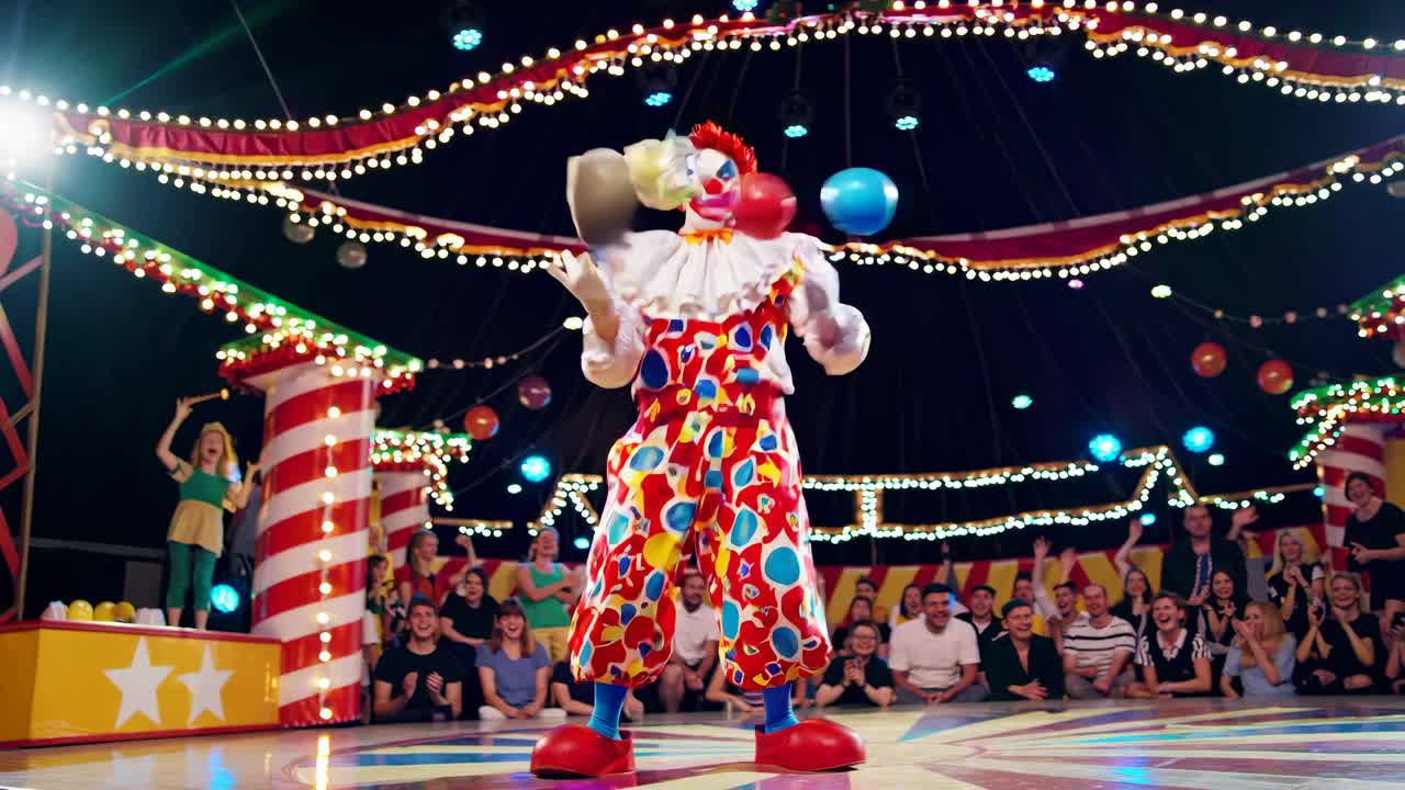 Wide-angle video shot of a colorful clown juggling under bright circus lights, with an audience