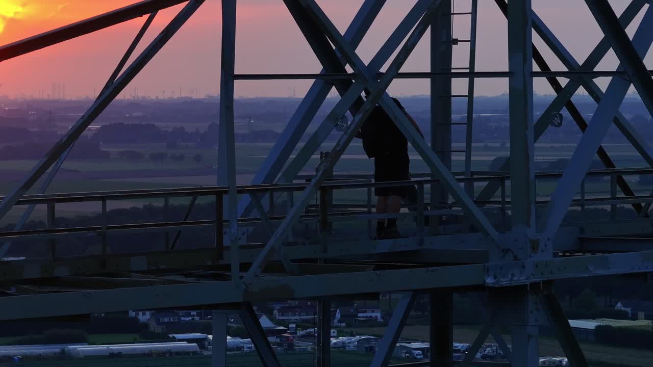 Man climbing up a steel structure at sunset in preparation for a base jump, with a rural backdrop