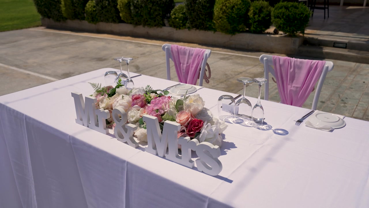 El video muestra una mesa de boda preparada al aire libre con un mantel blanco. Hay flores en el centro, copas y sillas con fundas. El letrero de 'Sr. y Sra.' complementa la decoración