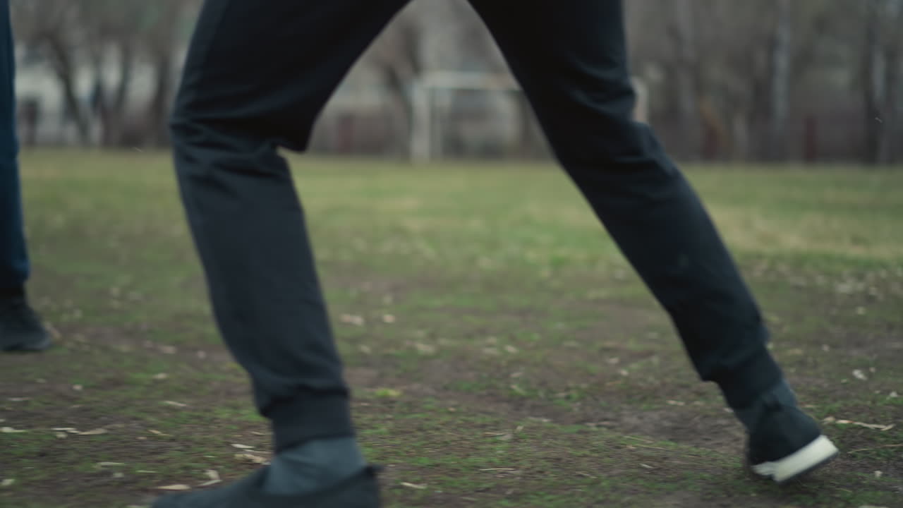 Close-up of two people engaged in a football game on a grassy field, while the background is blurred, showing trees, a goalpost, and another person behind them