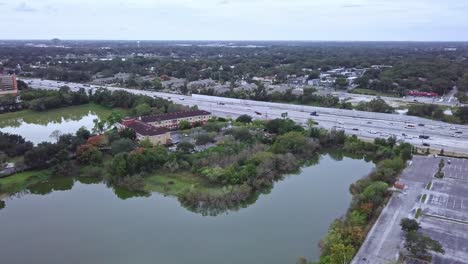 Forward-moving,-slow-rotating-aerial-drone-shot-of-busy-highway-showing-vehicular-traffic-with-small-lake-and-shopping-mall-parking-lot-in-foreground-and-buildings-and-landscape-in-background