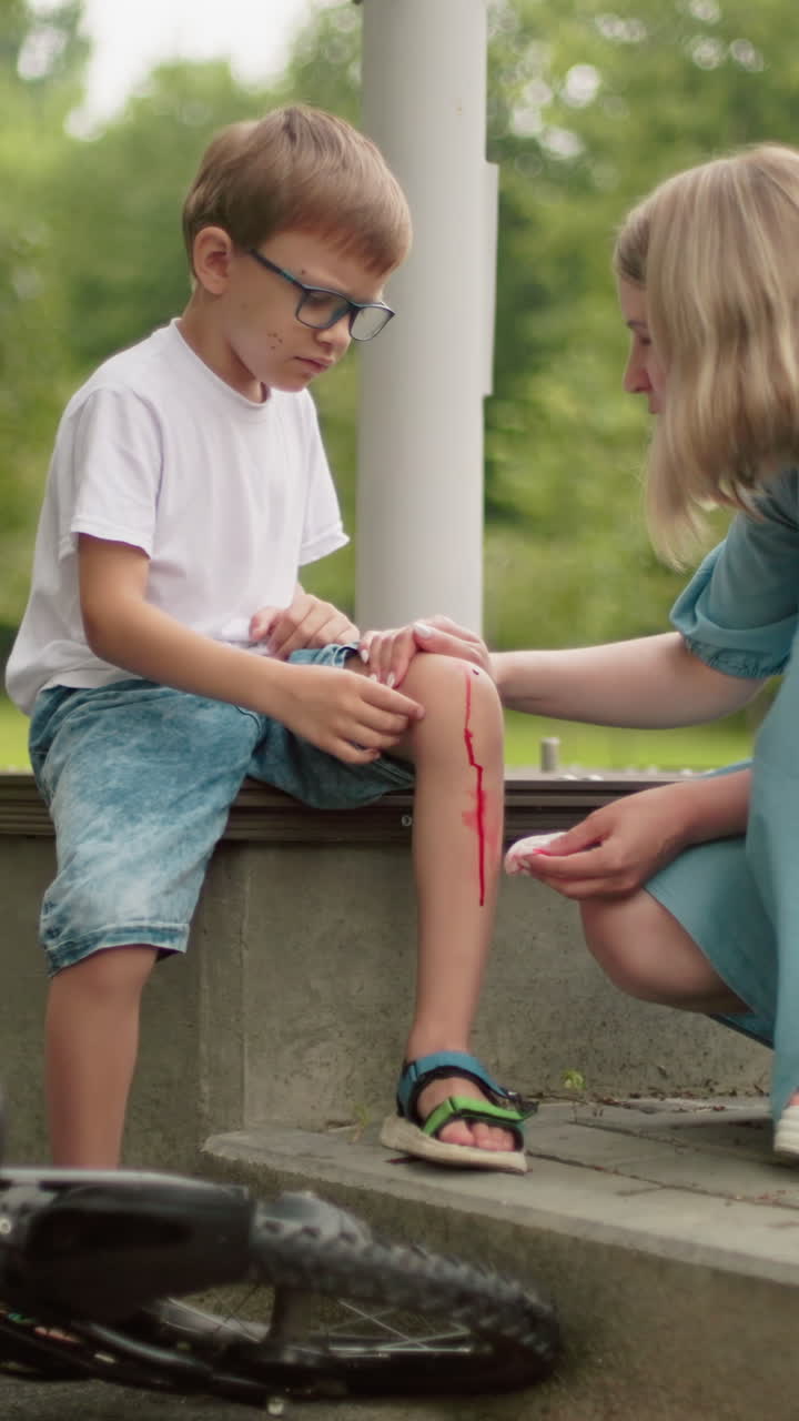 una madre se arrodilla frente a su hijo, limpiando una herida sangrienta en su rodilla con un pañuelo, el niño se sienta en un escalón, mirando infeliz