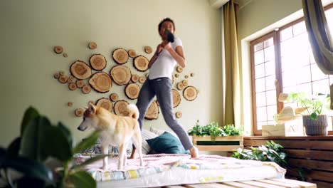 cheerful female student is dancing on bed singing in blow dryer having fun listening to music while its clever dog is standing on bed looking around.