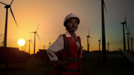 side view of asian female engineer in a helmet looking around while standing with arms akimbo in front of wind turbines rotating at sunset