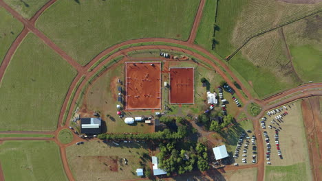 a wide top-down view of an equestrian sports area featuring a quadratic arena with red sand.