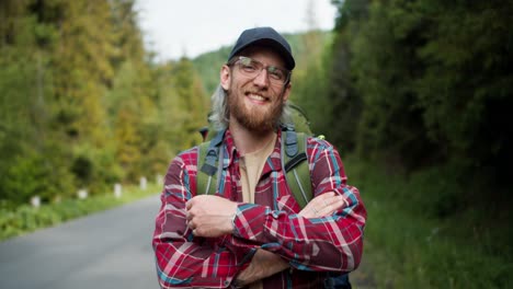 A-blond-man-tourist-with-a-beard-in-glasses-in-a-cap-stands-in-a-plaid-red-shirt-near-the-road-against-the-backdrop-of-the-forest