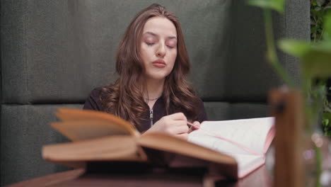 lady seated in soft green booth opens large brown book while glancing at another pink book on table, surrounded by indoor plants and simple decoration in calm cozy reading environment