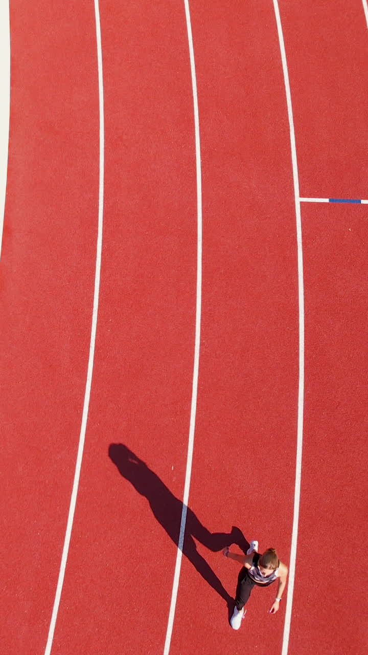 Aerial vertical shot of a female athlete sprinting on a red running track at a stadium, perfect for fitness and sports marketing