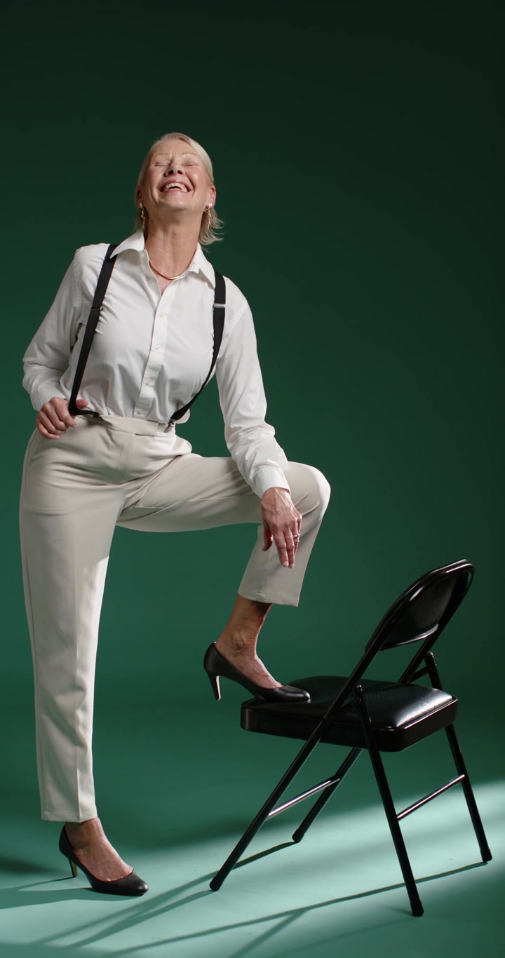 Vertical video: Senior woman placing foot on chair, modeling, pulling suspenders, smiling in studio