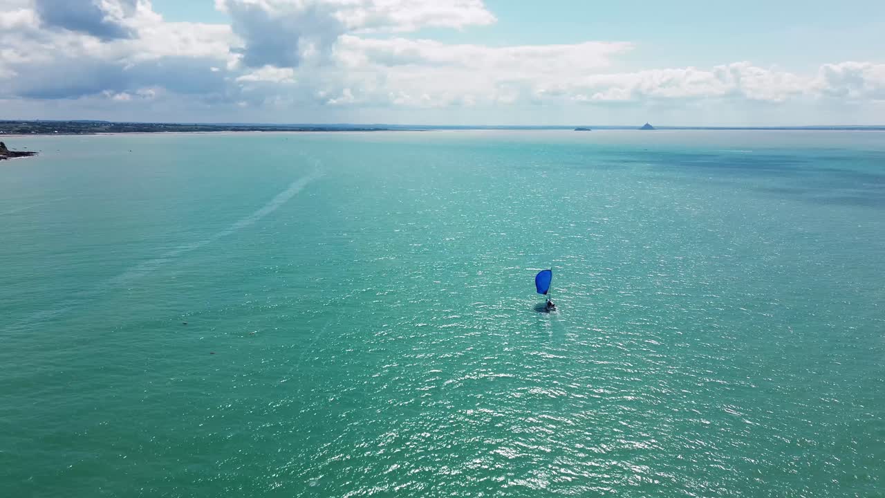 Drone footage follows a sailboat sailing across the coastal waters of Normandy, with Mont Saint-Michel appearing slowly on the horizon