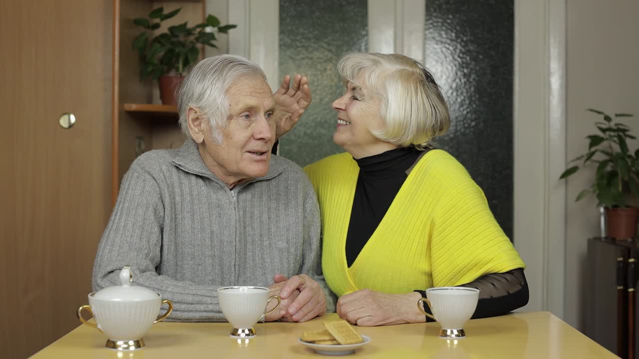 Happy old grandparents couple sit on table at home enjoy drinking tea together