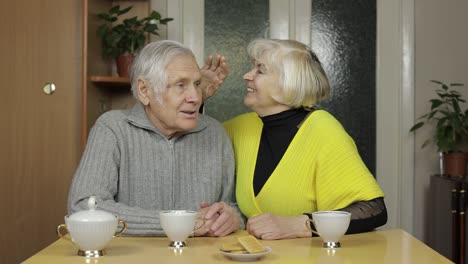 Happy-old-grandparents-couple-sit-on-table-at-home-enjoy-drinking-tea-together