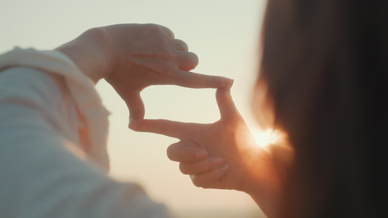 Close up rear view of woman framing sun between fingers, warm golden sunlight streaming through hands with lens flare and blurred river valley