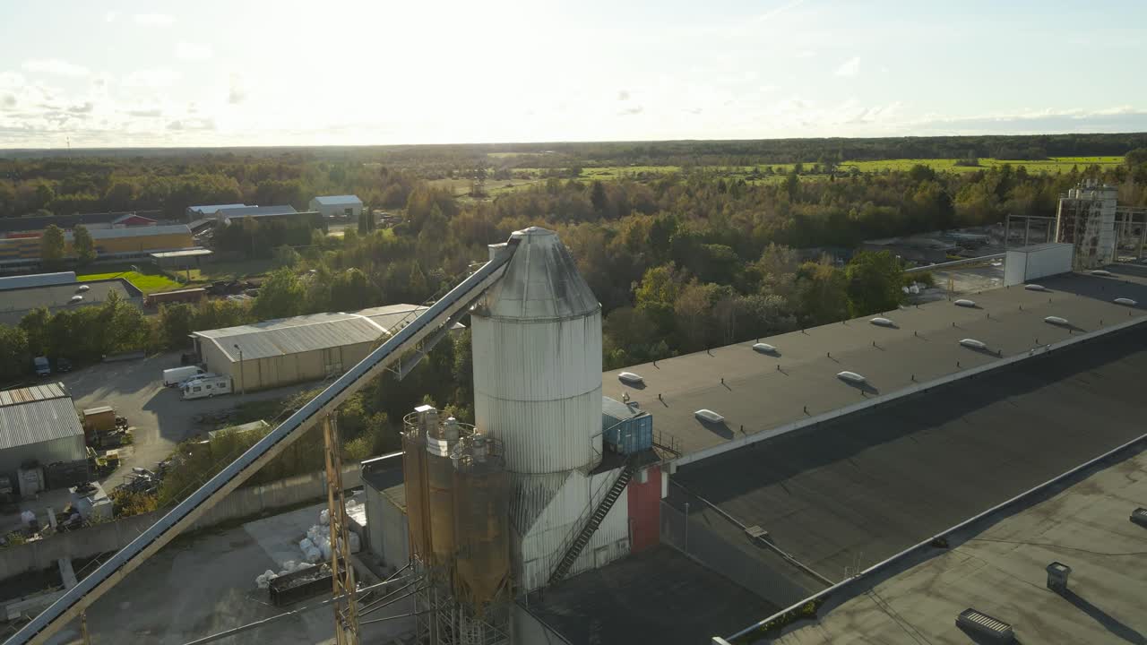 Aerial drone footage spinning around and orbiting a modern and tall concrete factory tower with pipes leading down from the structure to transport concrete to trucks in the middle on sunny countryside