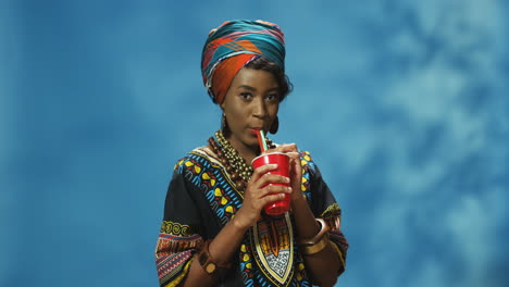 african american young woman in turban and traditional clothes drinking juice or soda and smiling at camera