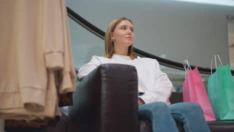 dama sentada en una silla de cuero en un centro comercial moderno con bolsas de compras de colores a su lado, vestida con una camiseta blanca casual y vaqueros, con una expresión enfocada, rodeada de exhibiciones de moda elegante