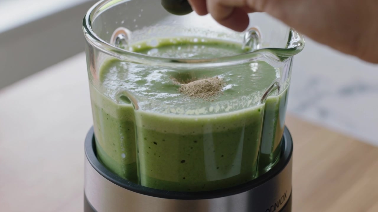 Una mano aggiunge con un cucchiaio della polvere a un frullato verde in un boccale di frullatore di vetro per scopi nutrizionali.
