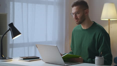 Der-Junge-Mann-Macht-Sich-Während-Einer-Online-Videokonferenz-Notizen-Mit-Kollegen,-Die-Von-Zu-Hause-Aus-Arbeiten-Und-Mit-Ihrem-Laptop-Am-Tisch-Sitzen,-Während-Sie-Remote-Arbeiten-Und-An-Einem-Geschäftstreffen-Teilnehmen