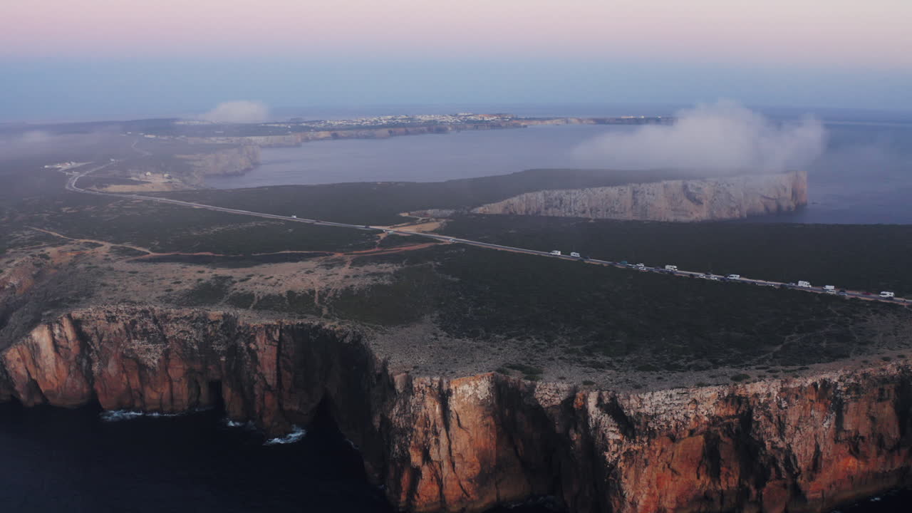 Dramatic aerial shot of rugged coastal cliffs with ocean waves below. Mist over a distant peninsula, connected by a scenic road at sunset. Sagres, Algarve, Portugal