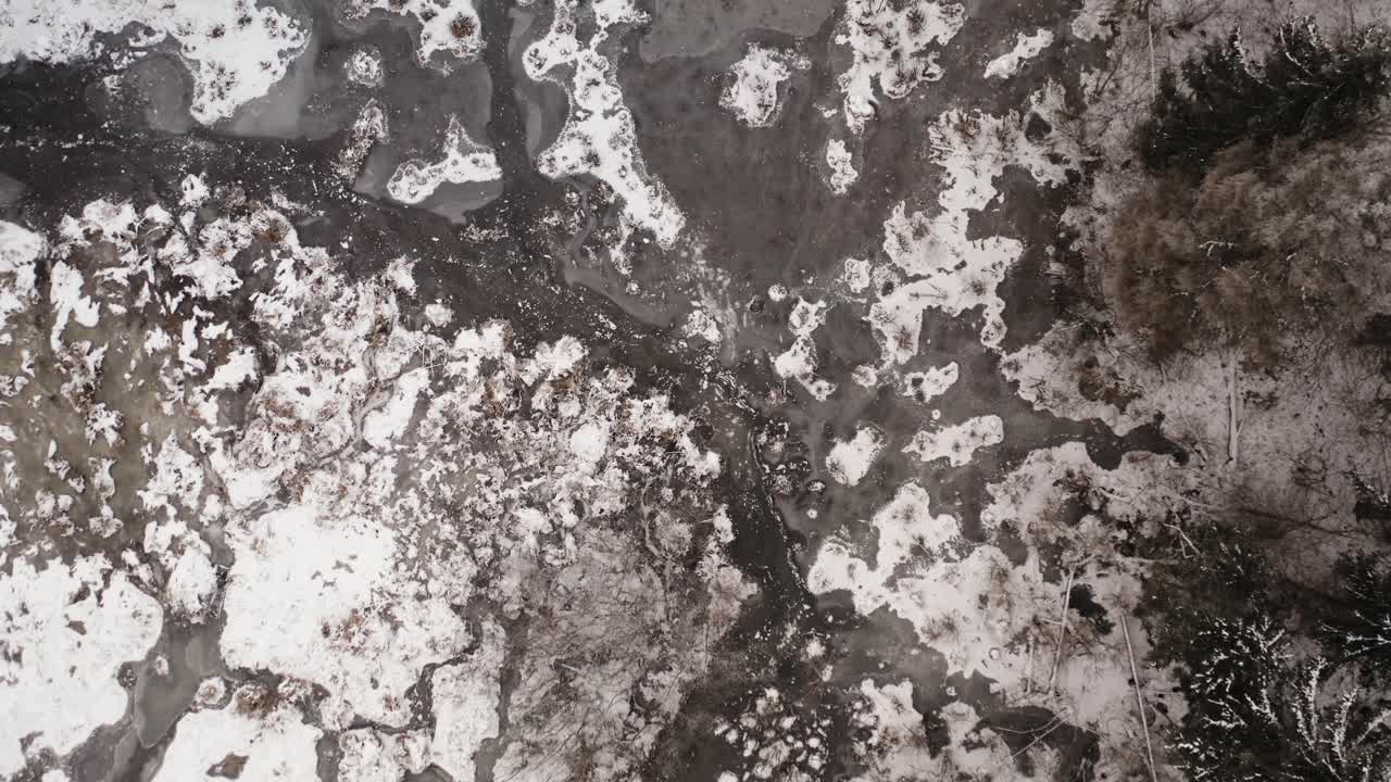 Aerial drone view of a partially frozen swamp in winter with icy terrain and snow covered landscape.