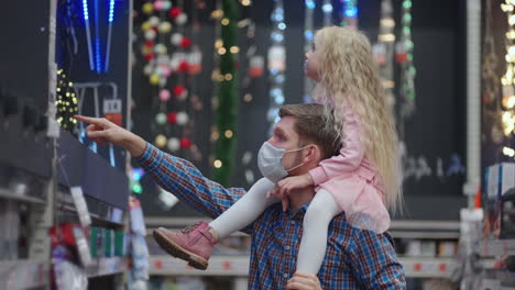 un padre y una hija sentados sobre sus hombros en un centro comercial eligen una guirnalda para la casa y un árbol de navidad para navidad durante la pandemia.