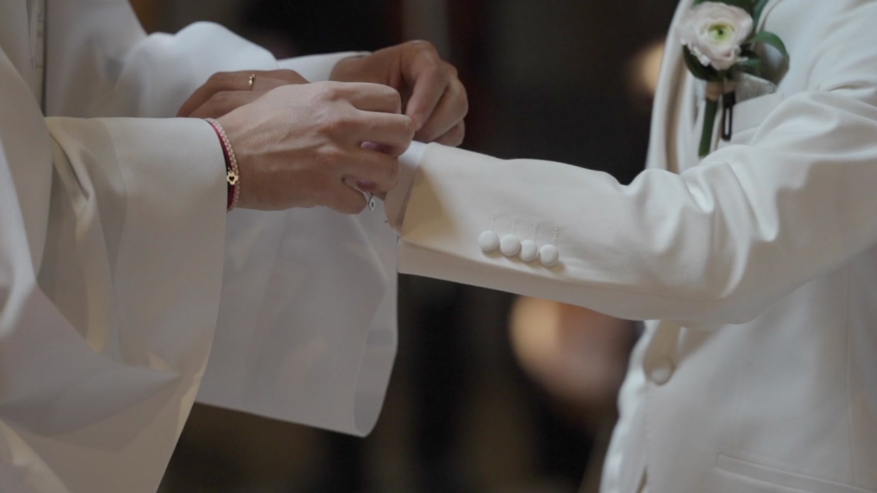 Close view of hands tying bracelet on wrist during wedding ceremony with white clothing and boutonniere visible
